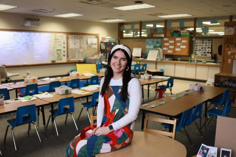 A cheerful young woman with long dark hair and a white headband smiles while sitting on a wooden table. She wears a vibrant, geometric-patterned overall. She is in a tidy classroom with small tables and blue chairs.