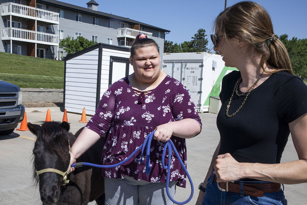 An Independence Academy student smiles with another woman as she leads a pony to a petting area. They are standing in a parking lot with an apartment complex in the background.
