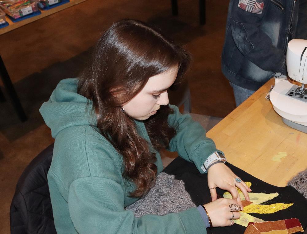 Bay High Focus Program student Zoey works with a patchwork sweater during the school's fashion club. She is using both hands to sew fabric in the shape of a sunflower onto a sweater. She has long hair and is wearing a sweatshirt and several rings on her fingers.