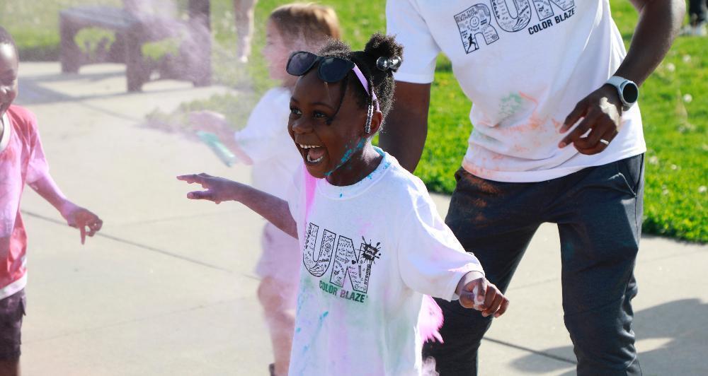 A girl, wearing a "RUN Color Blaze" shirt, joyfully runs through colored powder, during a color run.