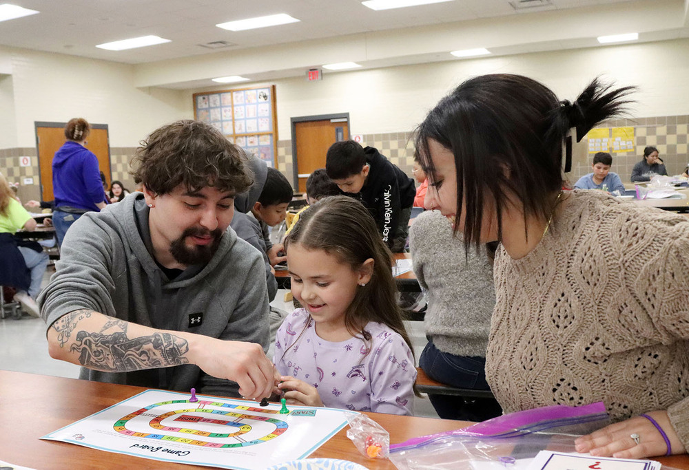 Two parents play a board game with their daughter in Everett Elementary School's cafeteria. They are moving game pieces across a board that contains a winding path. All three are smiling while they are sitting down at a cafeteria table.