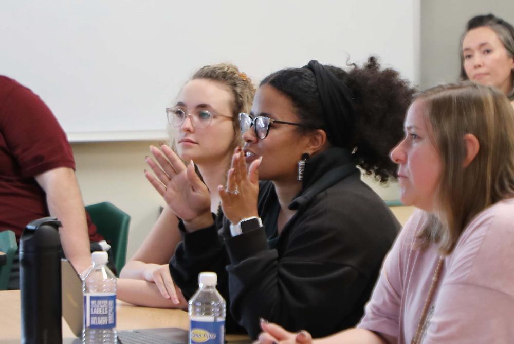 Four Lincoln Public Schools teachers participate in a class on the first night of the redesigned LPS ElevatED tenure-track program. They are taking professional learning classes at Southwest High School. There are three teachers sitting in a row at a table and a fourth teacher is sitting at another table in the background. The teacher in the middle is talking and has her hands spread apart to make a point about what she is saying.