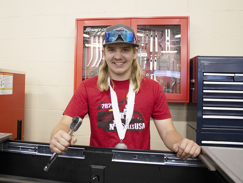 Northeast student Jaxson stands in front of a large toolkit that is mounted on a wall in a glass case. He is holding a wrench in his right hand and is standing in front of a storage bin in an industrial arts classroom.