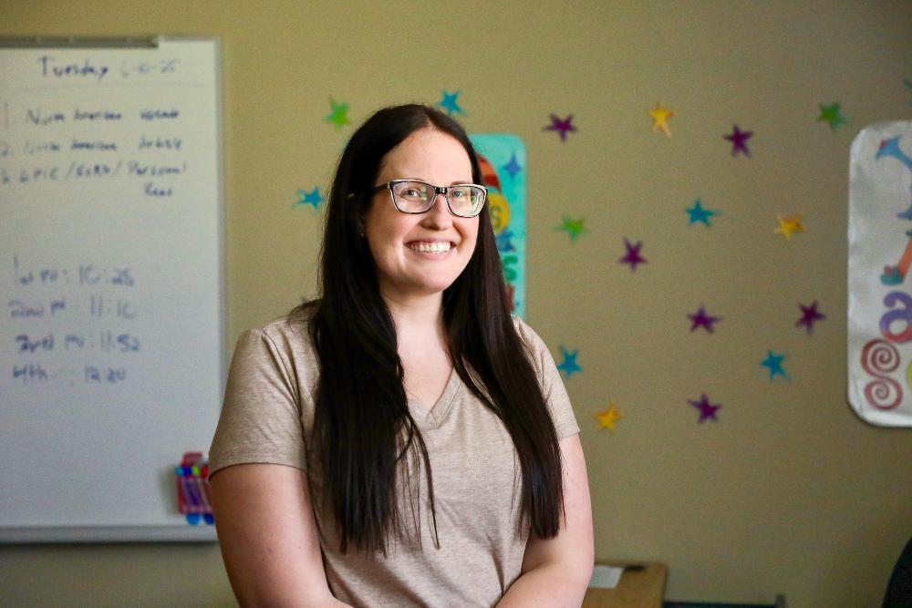 A woman with long dark hair and glasses smiles in a classroom. A whiteboard displays a schedule; the wall is decorated with colorful stars.