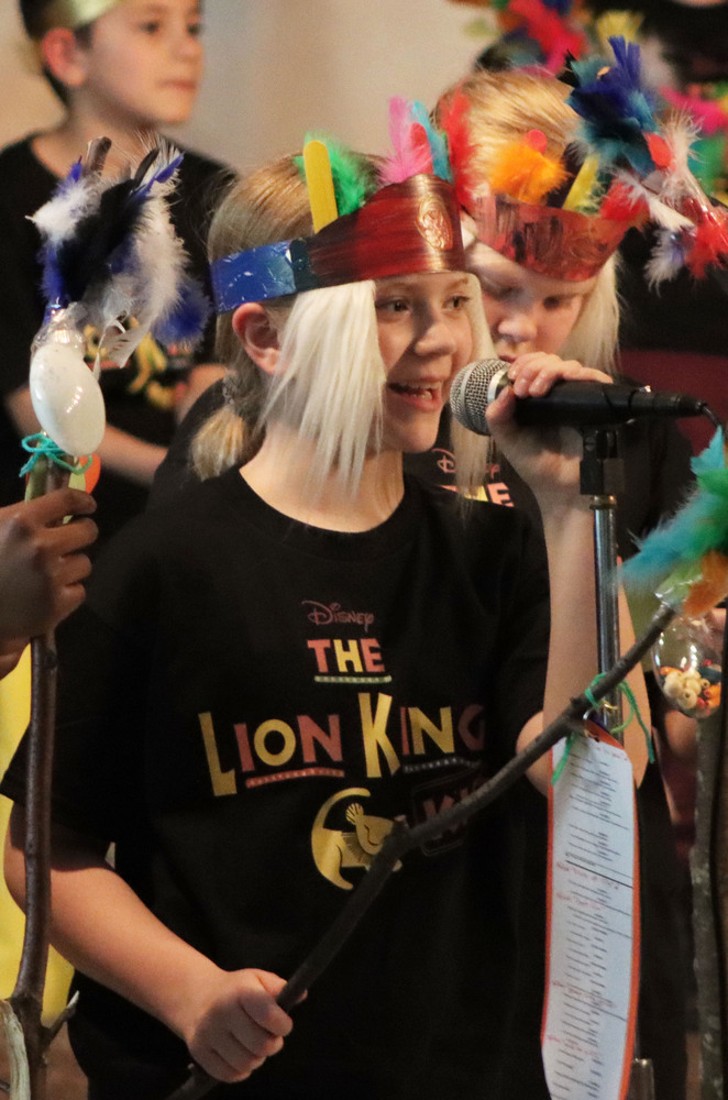 A Calvert Elementary School student smiles as she performs a song during The Lion King on stage. She is holding a microphone with her left hand.