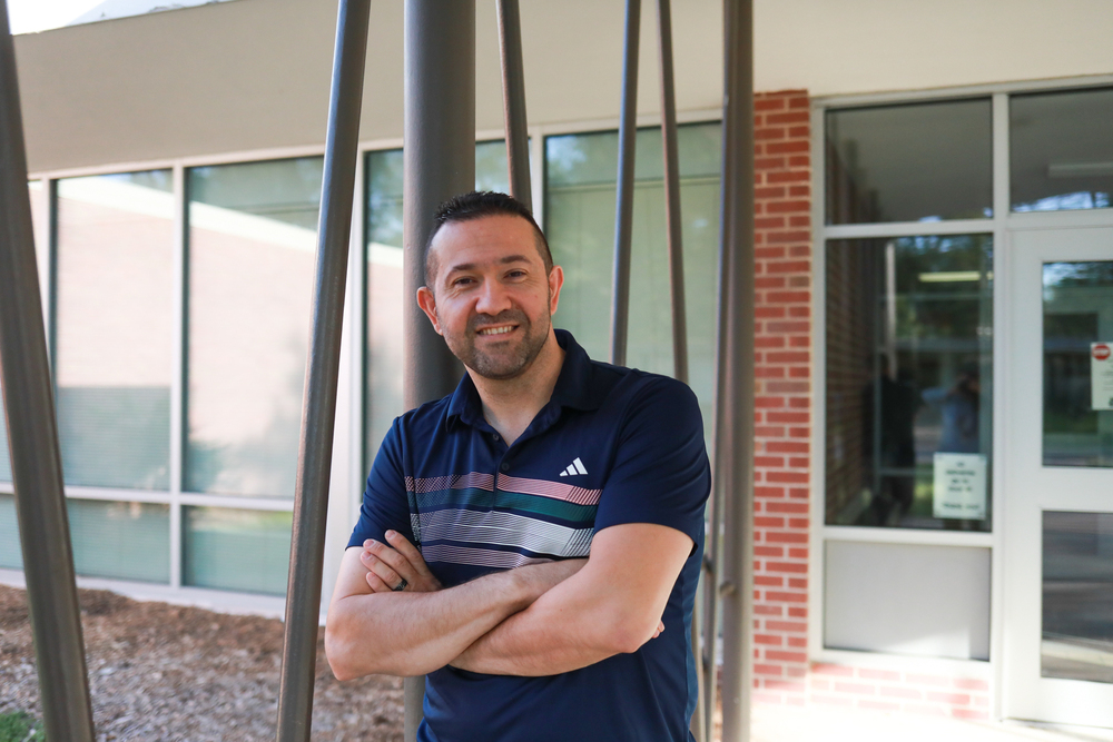 A smiling man wearing a navy blue Adidas polo shirt stands with his arms crossed outside a brick-and-glass building.