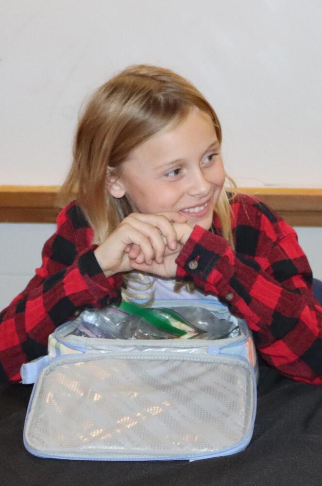 Rousseau Elementary School student Everly smiles at a table in a meeting room at school. She is looking to the right and has her hands clasped together above her lunch container. She is wearing a long-sleeved flannel shirt.