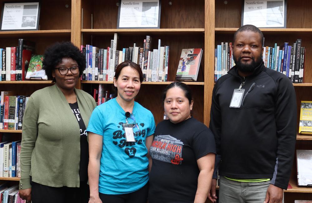 Four individuals stand before bookshelves, two women in the center, flanked by a woman and a man. They are likely library staff. The signs above read: "Nonfiction 040-050," "Nonfiction 073-977," and "Nonfiction 973-977."