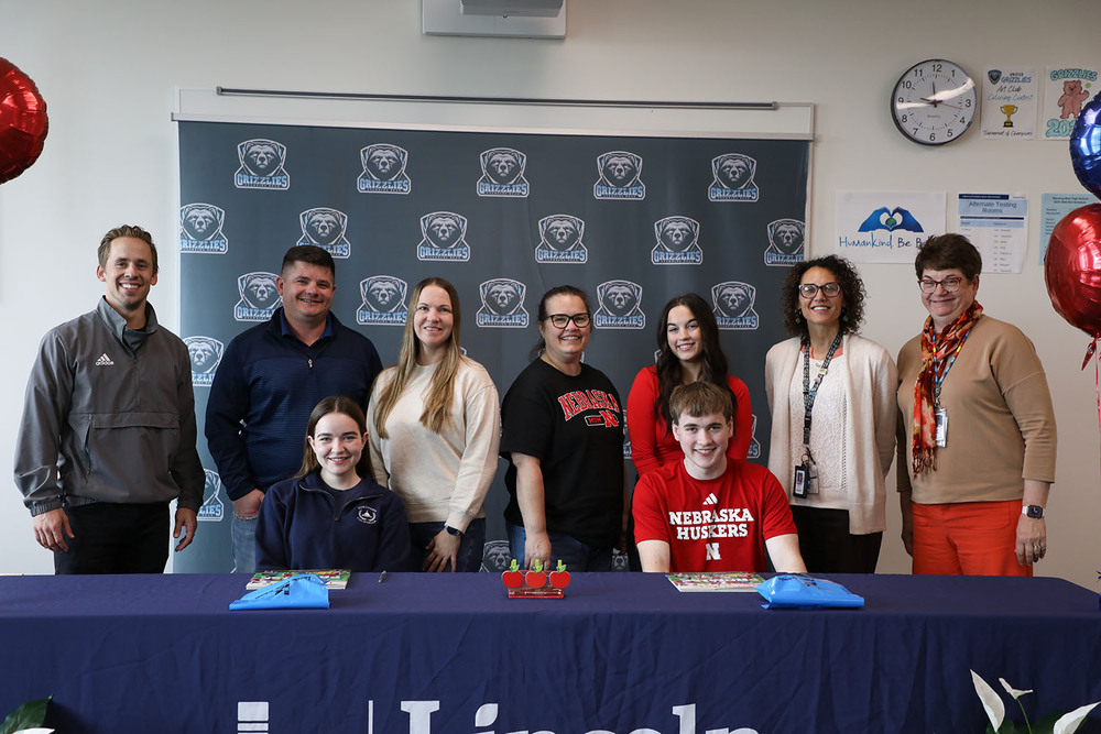 Two Standing Bear students smile as they sit at a table during a signing ceremony in a classroom. Seven parents, teachers and Standing Bear administrators are standing in a row behind them and smiling.