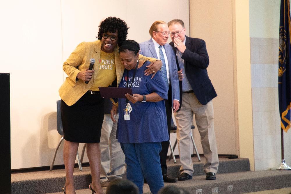 A woman in a yellow jacket presents an award to another woman wearing a blue shirt, inside what appears to be a school auditorium. The text on their shirts reads: "I ?? PUBLIC SCHOOLS".