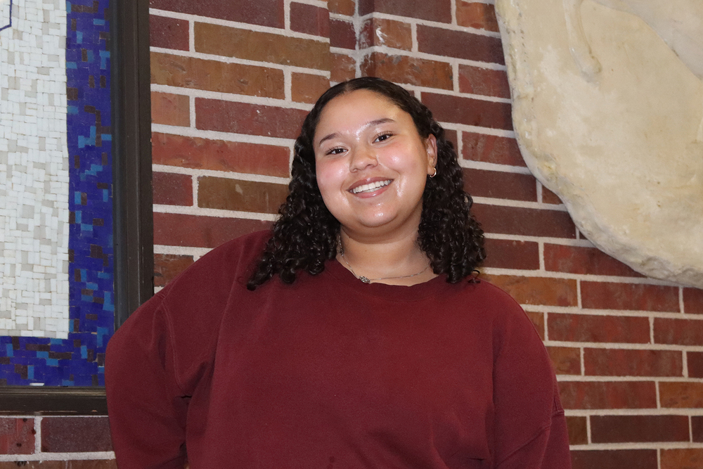 East High School senior Cyan smiles in front of a brick wall in a school hallway. There is a small portion of a mosaic that is hanging on the wall in the lefthand side of the photo.