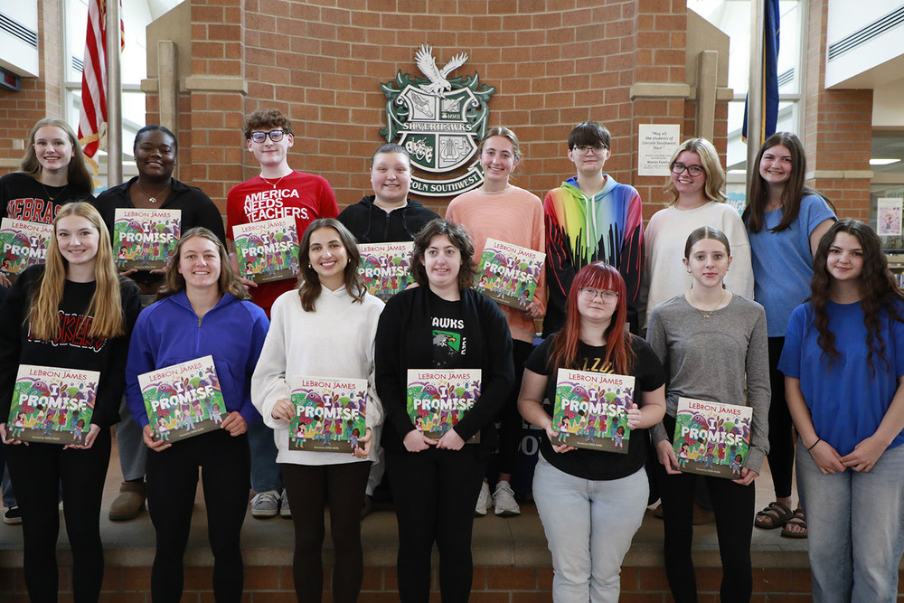 Fifteen students stand in a group, all holding the book "I Promise" by LeBron James. There is a brick wall with the Lincoln Southwest crest on it behind them.