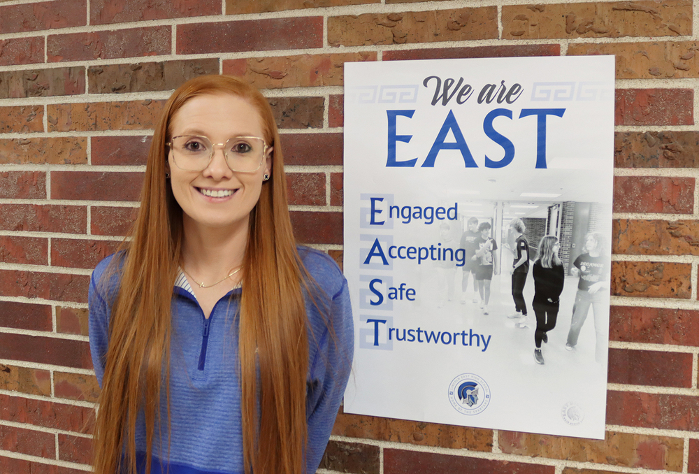 Sydney Austin smiles next to a poster that is attached to a brick wall in an East High School hallway. Austin is East's campus supervisor. The poster says, 'We are East: Engaged, Accepting, Safe, Trustworthy'