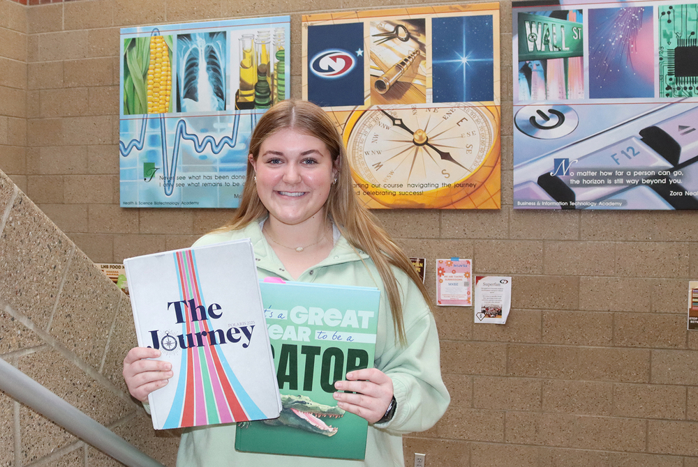 North Star senior Alexis smiles in a stairway of the high school. She is holding two yearbooks that she has worked on during her North Star journalism career. One cover says The Journey and the other says It's a great year to be a gator. There are several motivational signs on the wall behind her.
