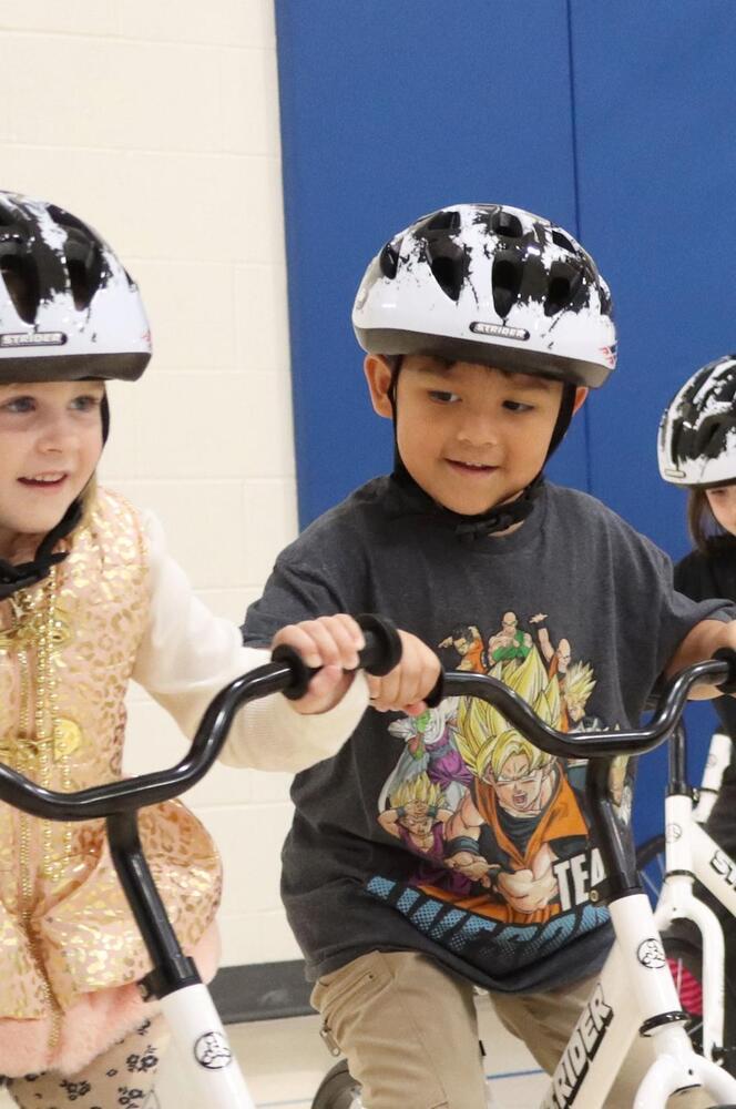 A Maxey Elementary School girl and boy are riding Strider bicycles in the school gym during a special presentation this fall. They are wearing helmets and have both hands on the handlebars of their bicycles. The girl is smiling and looking forward while riding her bicycle, and the boy is smiling and looking over at the girl while riding his bicycle.