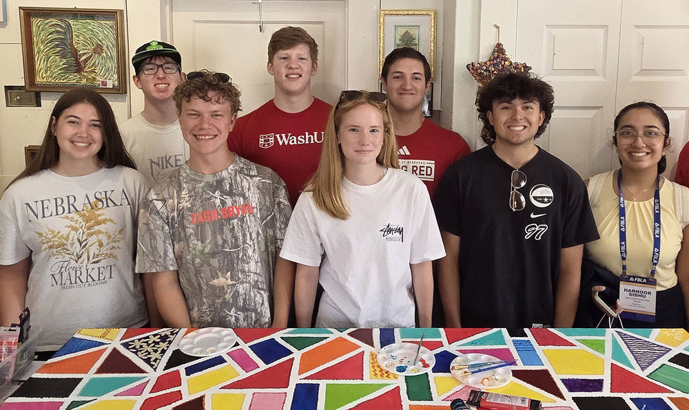 Eight Southwest High School students stand together in two rows. There are three girls and five boys in the photo. They are taking part in an artistic activity in a room that has several paintings on the back wall.
