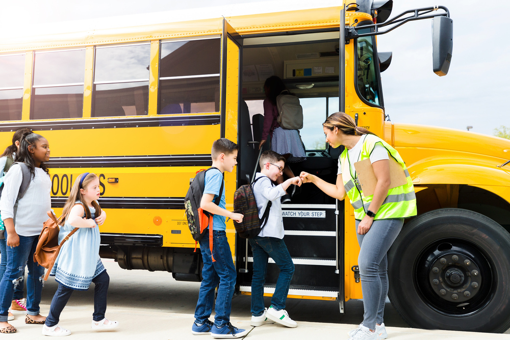 A young woman in a safety vest with a cliboard welcomes a line of students who are boarding a yellow school bus