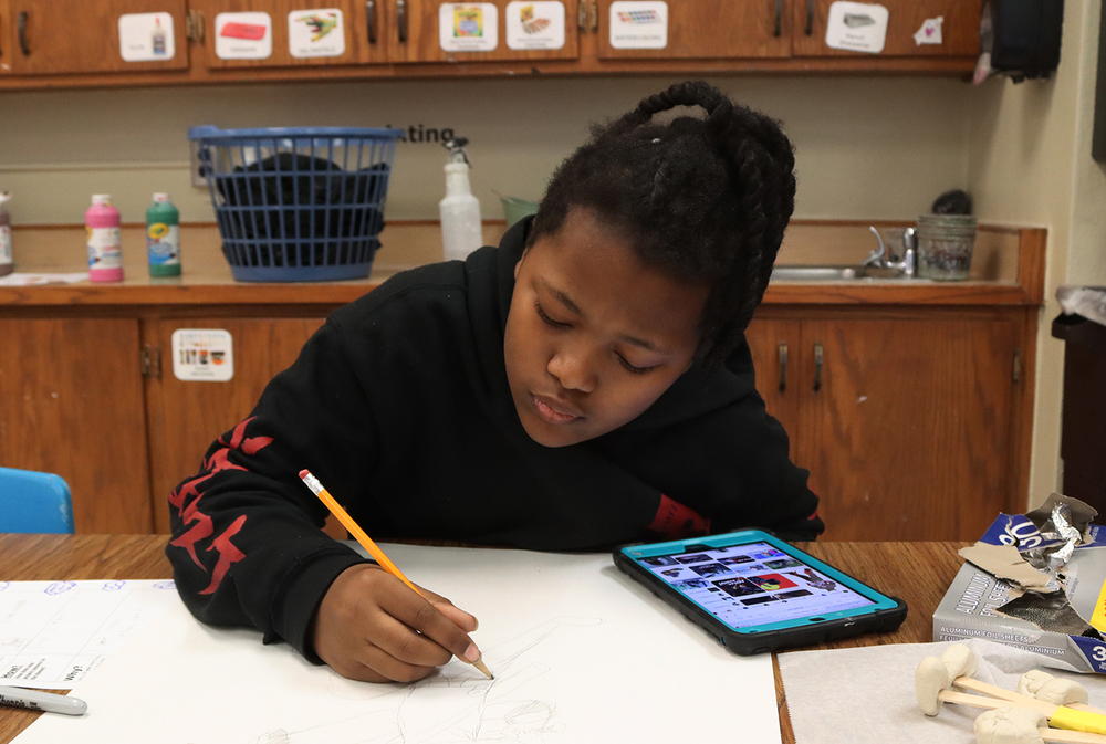 Saratoga Elementary School fifth grader Ka'Avontae uses a pencil to sketch an image of a ninja superhero during an art project in class. There is an iPad on the table next to him.