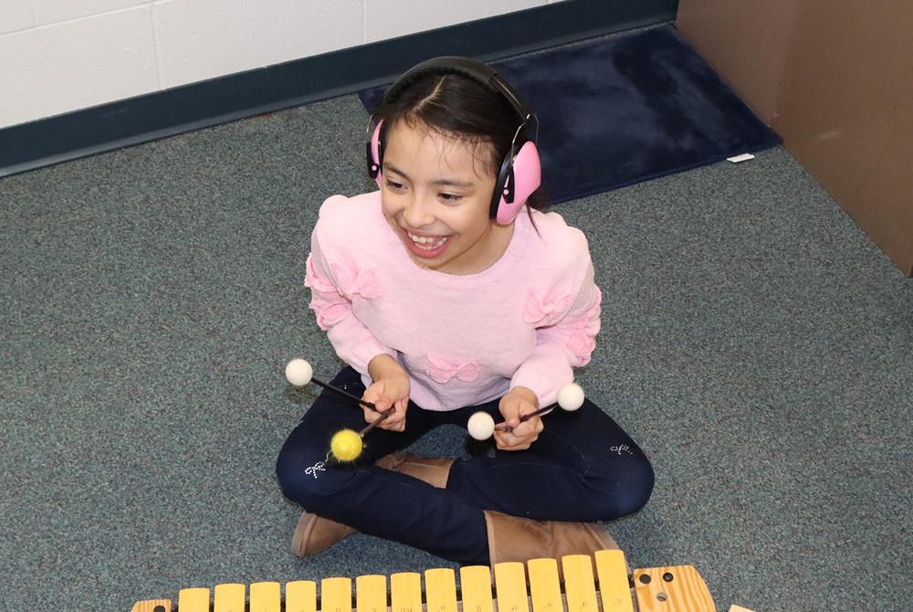 A Riley Elementary School student beams as she plays the xylophone in class. She is sitting down next to the instrument and is holding mallets in both hands.