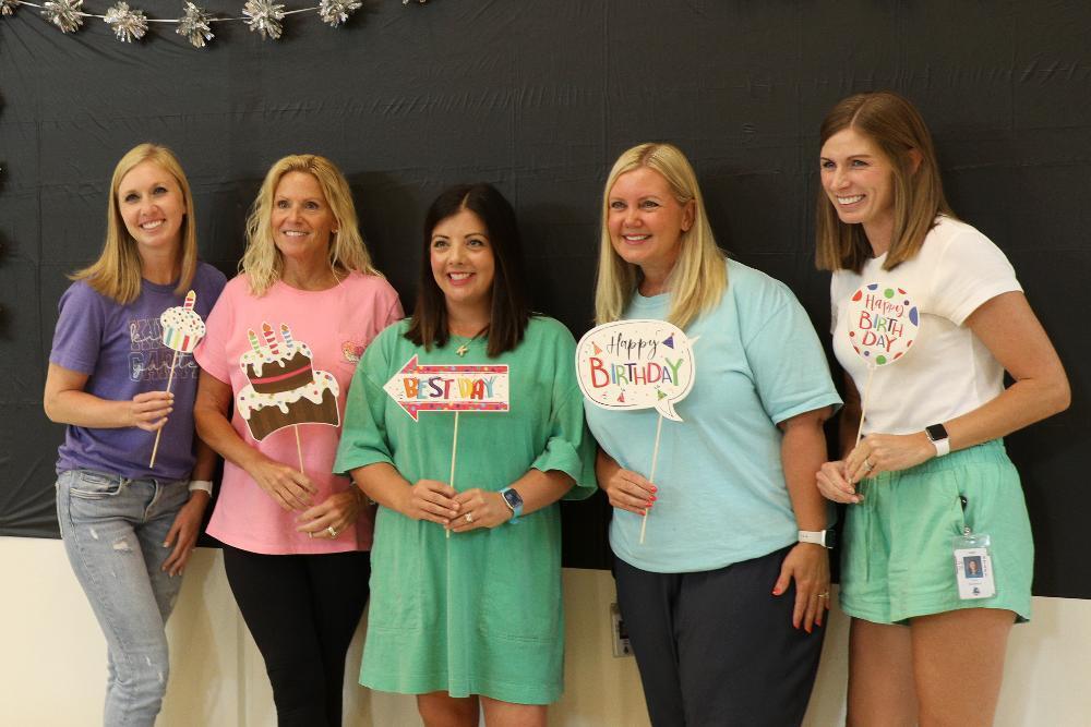 A row of five teachers smile toward a camera at Wysong Elementary School. From left, they are holding various items to celebrate the school's new Pack mentality program.