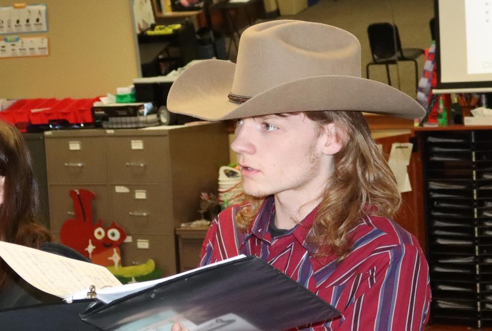 Northeast High School senior Tayler performs with the Chamber Singers group in the choir room. He is wearing a cowboy hat and is holding a songbook while he is singing.
