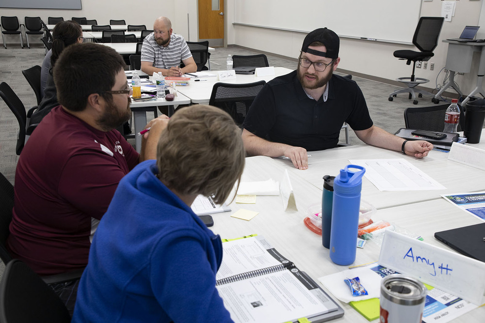 Three Lincoln Public Schools music staff are discussing a crowd management prompt during the summer 2026 NCS4 training. They are sitting together at a large table. There are several other people in the background.