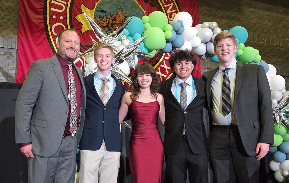 From left, Southwest High School business teacher Josh Hinrichs stands next to Cooper, Mylee, Xavier and Alex on a stage at Boston University. They took part in the National Junior Achievement Stock Market Challenge. They are wearing formal attire for an awards ceremony.