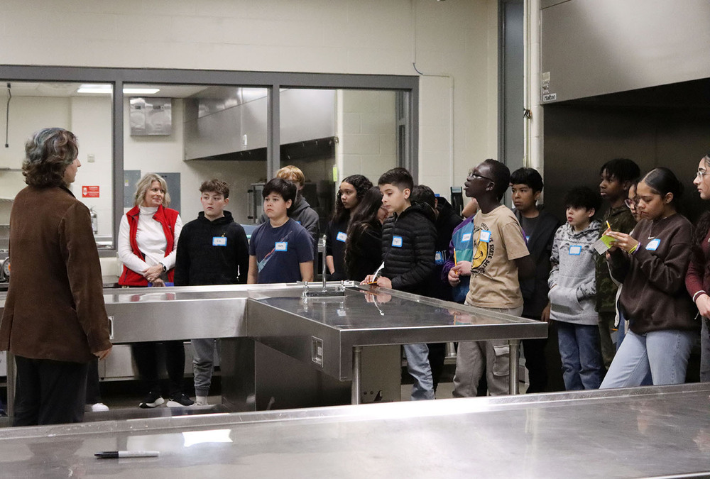 Thirteen middle school students listen to a presentation in the main culinary arts classroom at The Career Academy. A presenter is standing behind a metal kitchen counter at the far lefthand side.