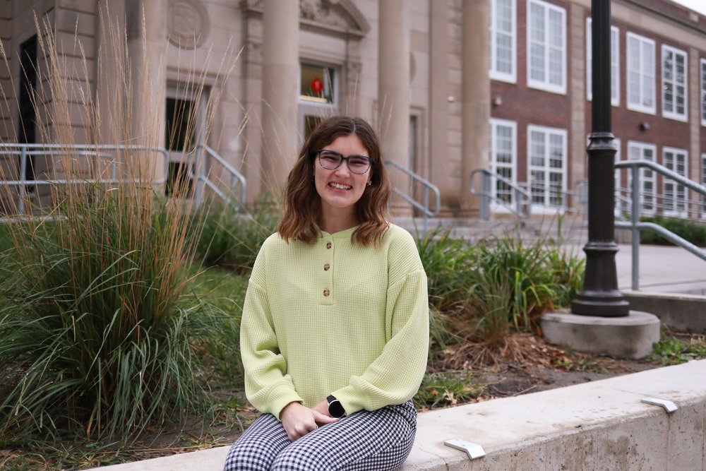 A person wearing glasses, a light yellow sweater and black-and-white checkered pants sits on a concrete ledge in front of a landscaped school building, smiling at the camera.