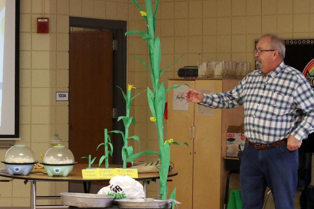 A man in a plaid shirt presents an educational display on a table. It features green paper corn stalks of different heights illustrating growth, and two clear popcorn makers filled with popcorn.