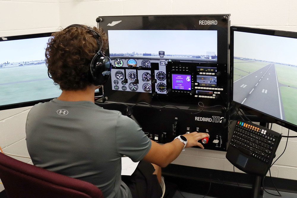 Aviation and Technical Education Focus Program student Samuel operates a flight simulator at North Star High School. He is sitting in front of three screens that are showing a simulated view of Lincoln from the cockpit of an airplane. He has his hands on cockpit instruments and controls.