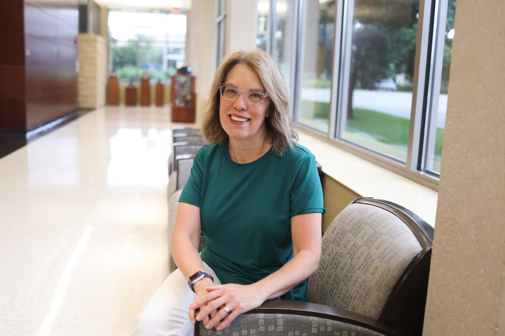 Smiling woman wearing glasses sits in a chair in a bright hallway with large windows.