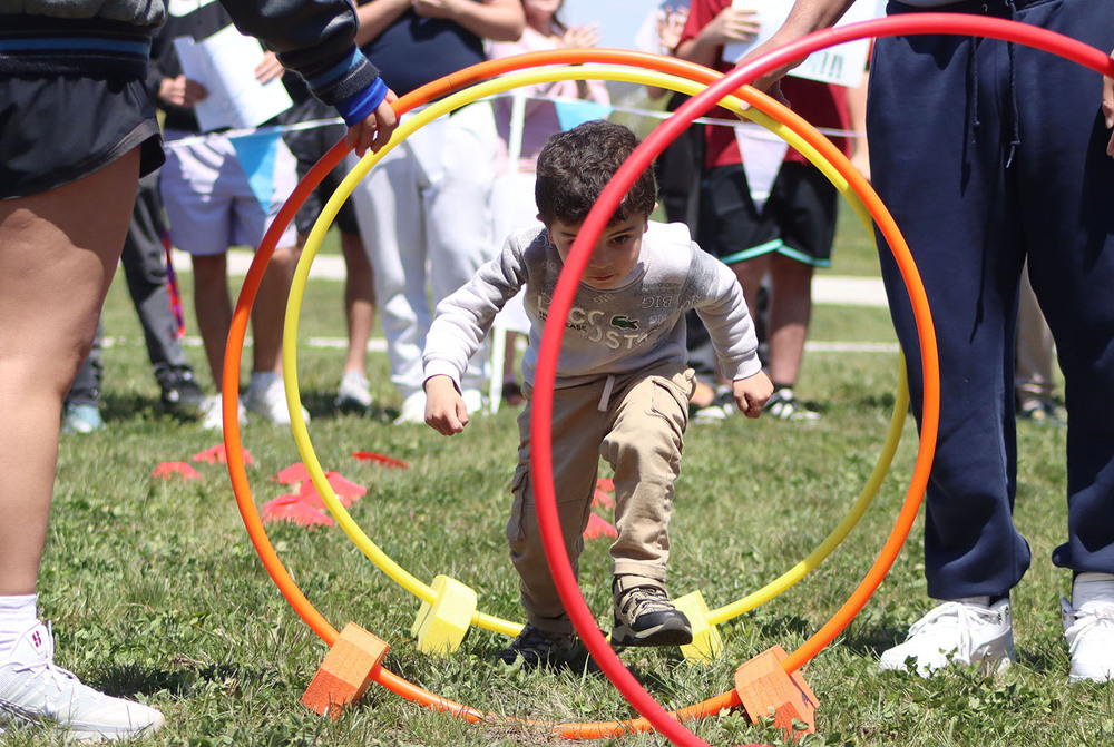 A preschool student moves his way through a series of three hula hoops at the inaugural Cubs Field Day. Two high school students are holding up the hoops for him to run through.