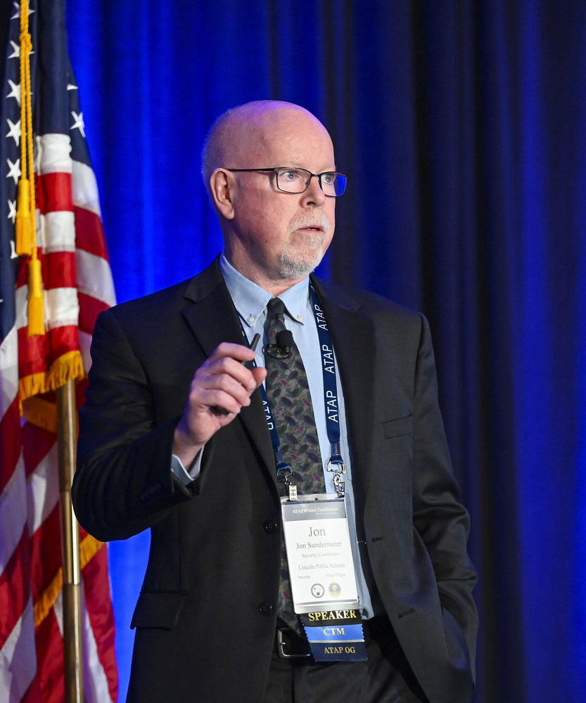 Jon Sundermeier speaks at a national conference. He is wearing a suit and tie and is facing left to right while standing on a stage.