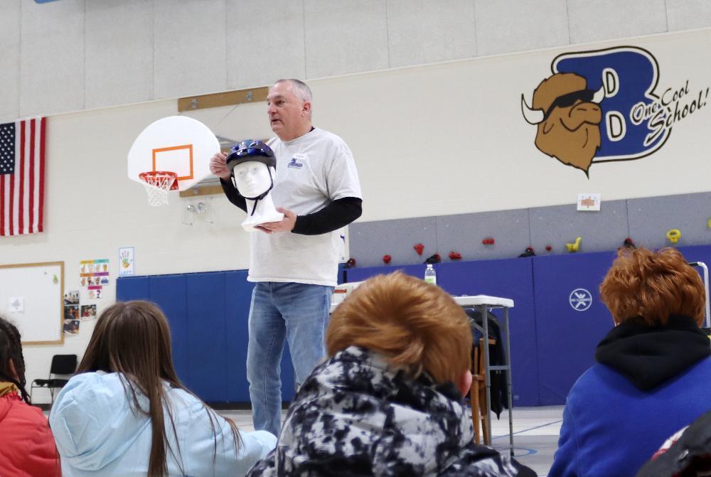 A man holds a blue bicycle helmet on a mannequin head, demonstrating to students sitting on a gym floor. An American flag hangs on the left, and a "BD One Cool School" buffalo logo is on the back wall.