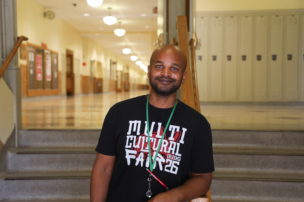A smiling LPS staff member sits on a staircase in a school hallway, wearing a “Multicultural Fair” T-shirt and green lanyard.