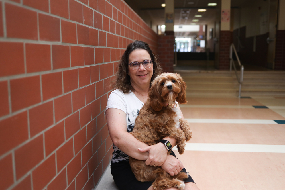 A woman smiles while holding a small brown therapy dog in a school hallway.