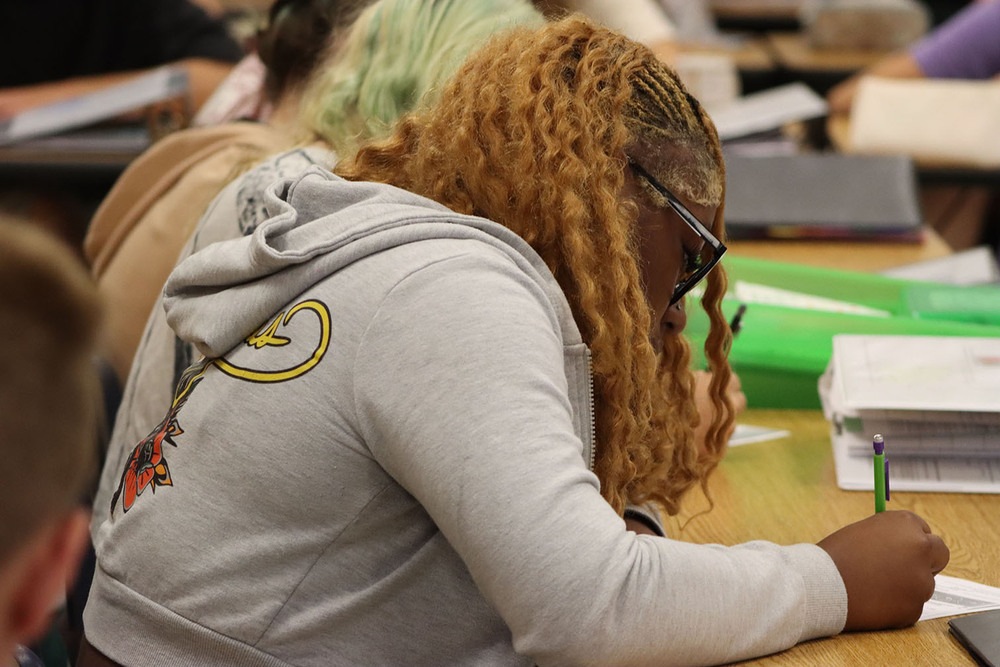 A World Experiences student takes notes midway through the class. She is facing left to right and is writing with a mechanical pencil in her right hand.
