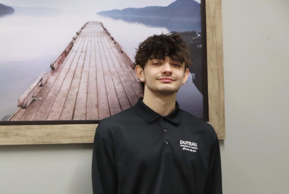 A young man stands before a picture of a wooden dock extending into a calm body of water. He wears a black polo shirt with "DUTEAU CHEVROLET BUICK Better by you" embroidered on it.
