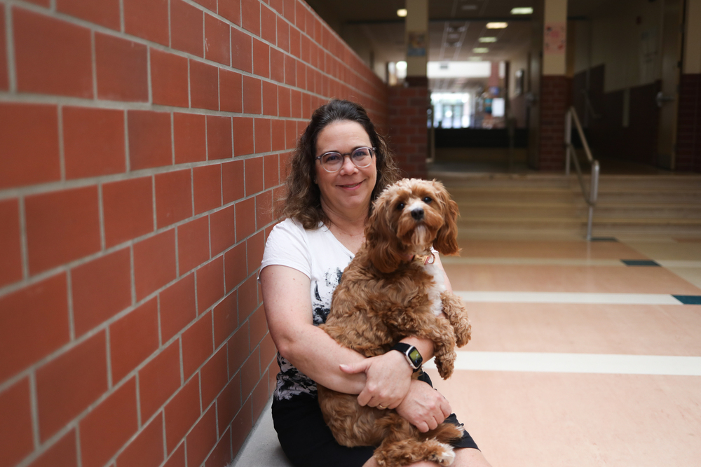 A woman smiles while holding a small brown therapy dog in a school hallway.