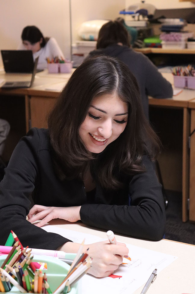 A North Star student smiles as she uses a marker to color in a portion of her fashion design drawing. She is using her right hand to fill in an image at a desk. Many pencils and markers are in a tray next to her on the table.