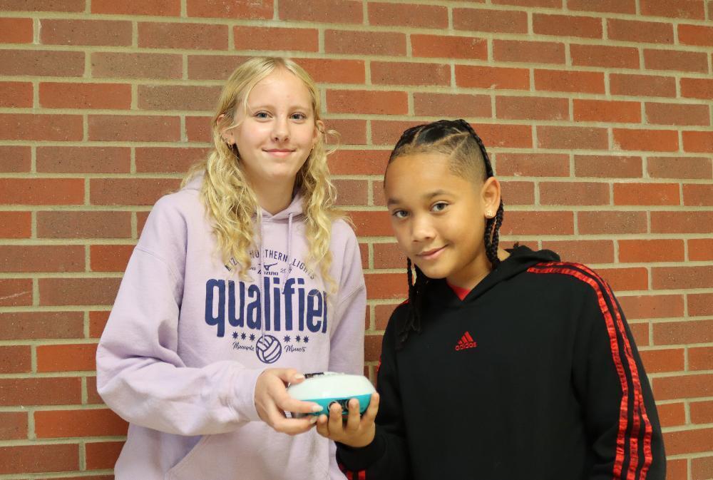 From left, Pound Middle School students Liz and MihKing hold a small robot that they used in their Math Course 2D class. Liz has long, curly hair and is holding the righthand side of the robot. MihKing is using his right hand to hold the lefthand side of the robot. They are standing in a hallway with a brick wall behind them.