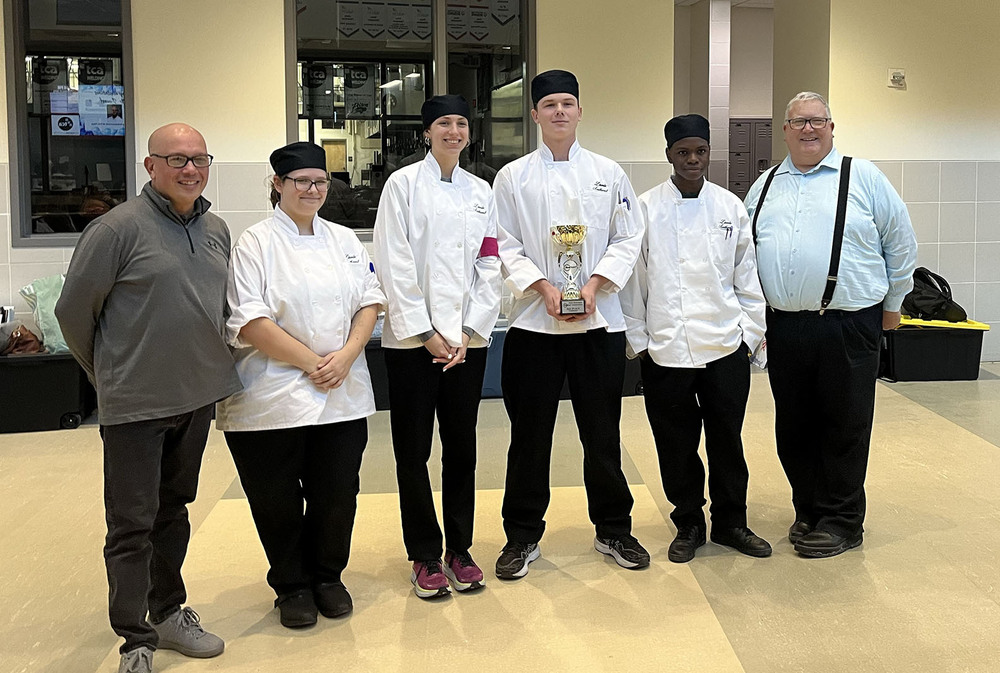 Four Southwest High School students stand in a row in between two adults. They are smiling with a state culinary contest trophy. A boy in the middle of the row is holding the vertical trophy.