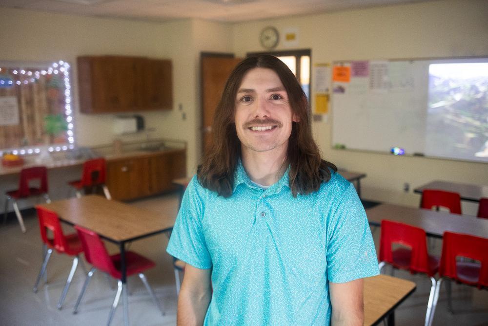 A man standing in a classroom wearing a cyan shirt.
