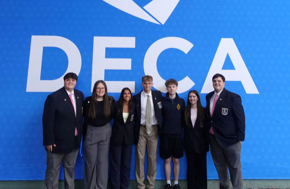 Seven Southeast High School students stand next to each other in a row at the DECA International Career Development Conference. The three girls and four boys are standing in front of a large wall that has the words DECA on it. They are wearing business attire for their competitive events.