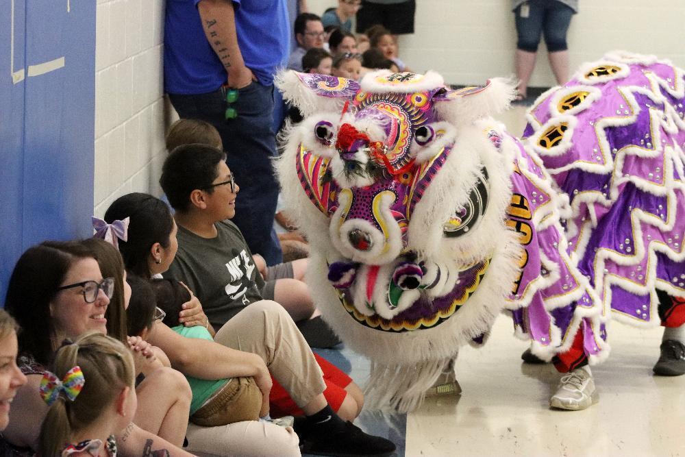 A lion dance costume performs for a seated audience in a gymnasium. The colorful costume is ornate, with purple and white accents.