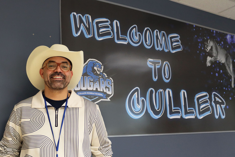 Culler Middle School Principal Daniel Church smiles in the main office. He is standing in front of a large poster that says "Welcome to Culler" with a picture of a Cougar on the right and the school logo on the left.