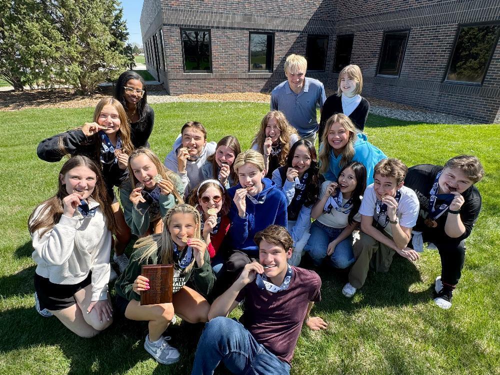 A large group of teenagers, many holding medals, are sitting on a grassy lawn outside a brick building. They appear happy and are possibly celebrating an accomplishment. One holds a trophy inscribed "UNIVERSITY NATIONAL QUALIFIER".