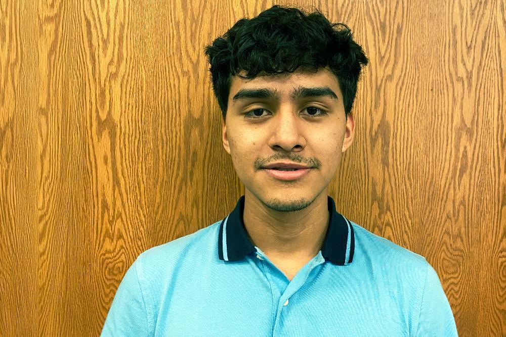 A young man with dark, curly hair poses against a wood-paneled wall. He wears a light blue collared shirt.