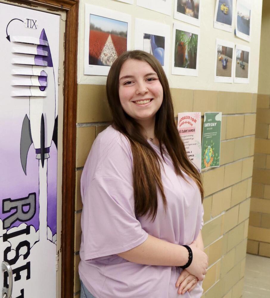 A young woman with long brown hair, wearing a lavender shirt, stands smiling against a wall beside a rocket-themed locker. A fundraiser poster and Earth Day notice are visible nearby.